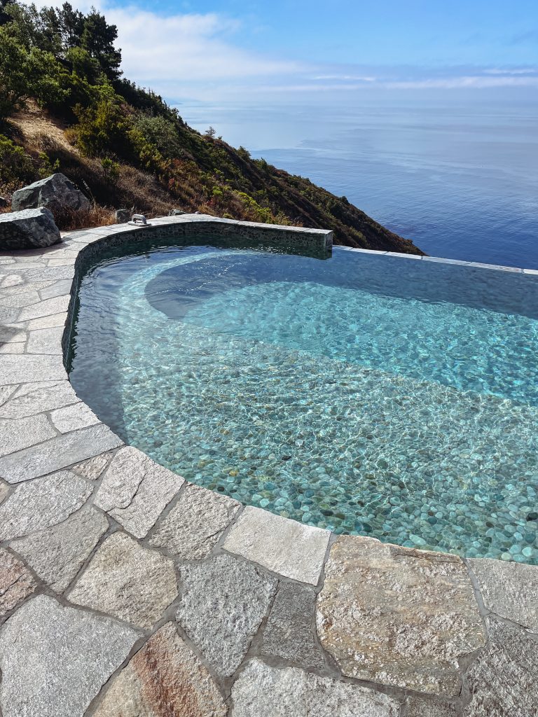a pool with a stone walkway and a hill and a body of water